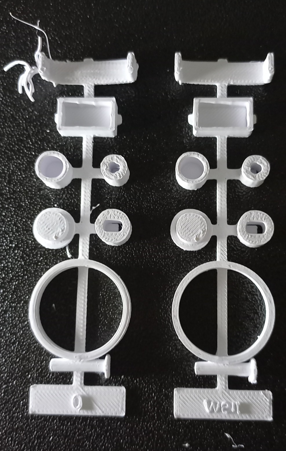 Photo of two white 3D printed fit-test sprues on a black surface, each holding bores, seats, a ring and a small bracket, one of them with stringy strands at the top