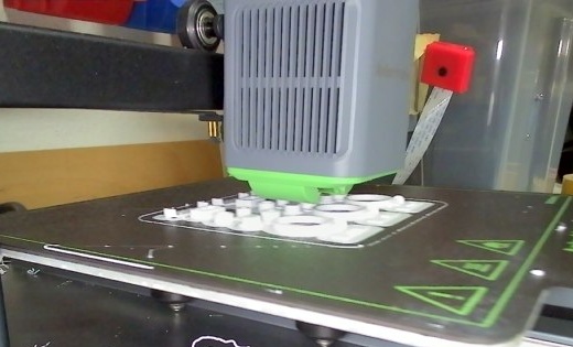 Webcam snapshot from inside a 3D printer: the print head moves over the build plate while the first layers of the small test parts are printed